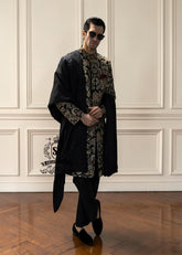 This luxurious royal black velvet sherwani, adorned with opulent gold hand embroidery, exudes a bold and dignified presence. The plush velvet fabric highlights the depth of the intricate hand embellishments, creating a truly regal style befitting a groom or a grand ceremony.