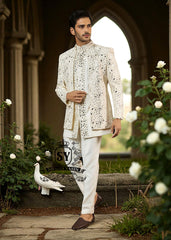 Man wearing a traditional white embroidered Prince  jacket and pants standing in a garden with flowers and a stone archway.