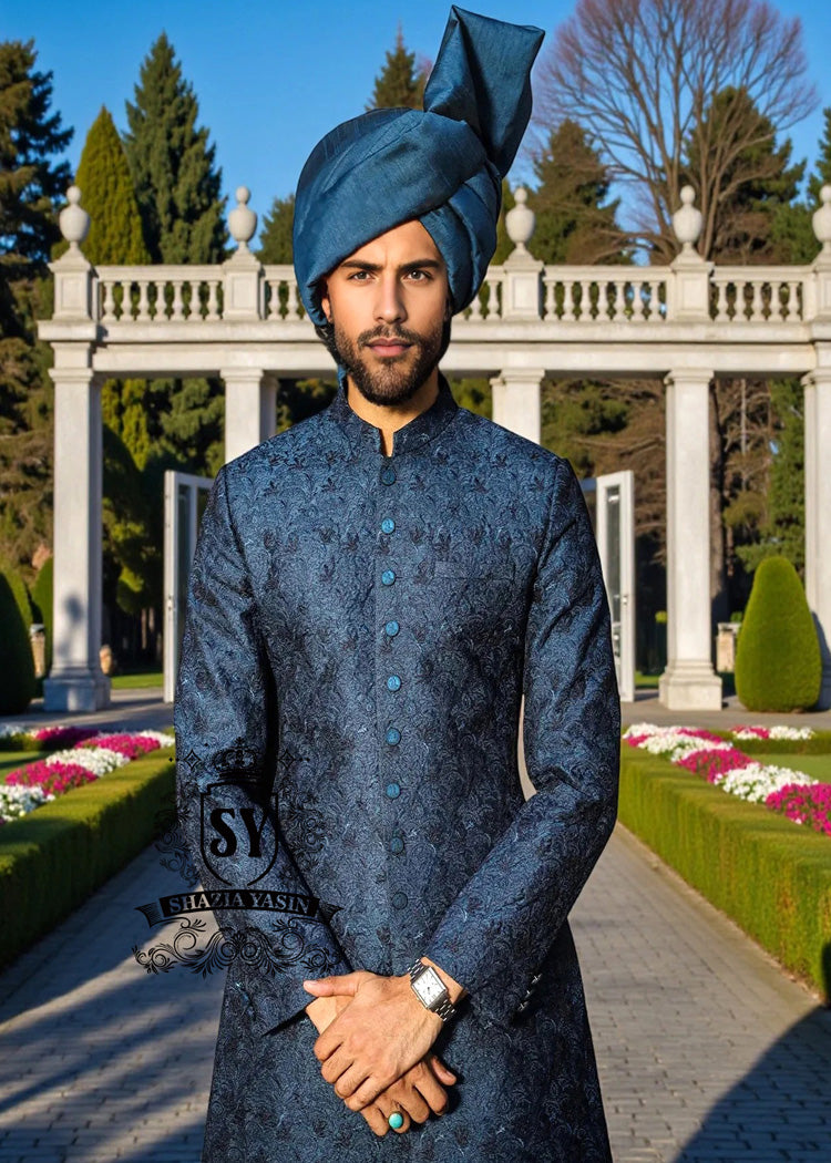 Man in a blue sherwani with a matching turban standing in a garden with white columns and greenery.