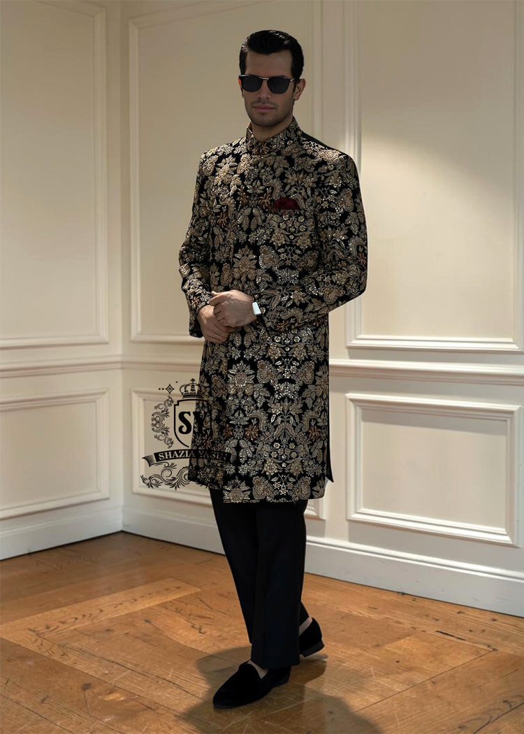 This luxurious royal black velvet sherwani, adorned with opulent gold hand embroidery, exudes a bold and dignified presence. The plush velvet fabric highlights the depth of the intricate hand embellishments, creating a truly regal style befitting a groom or a grand ceremony.